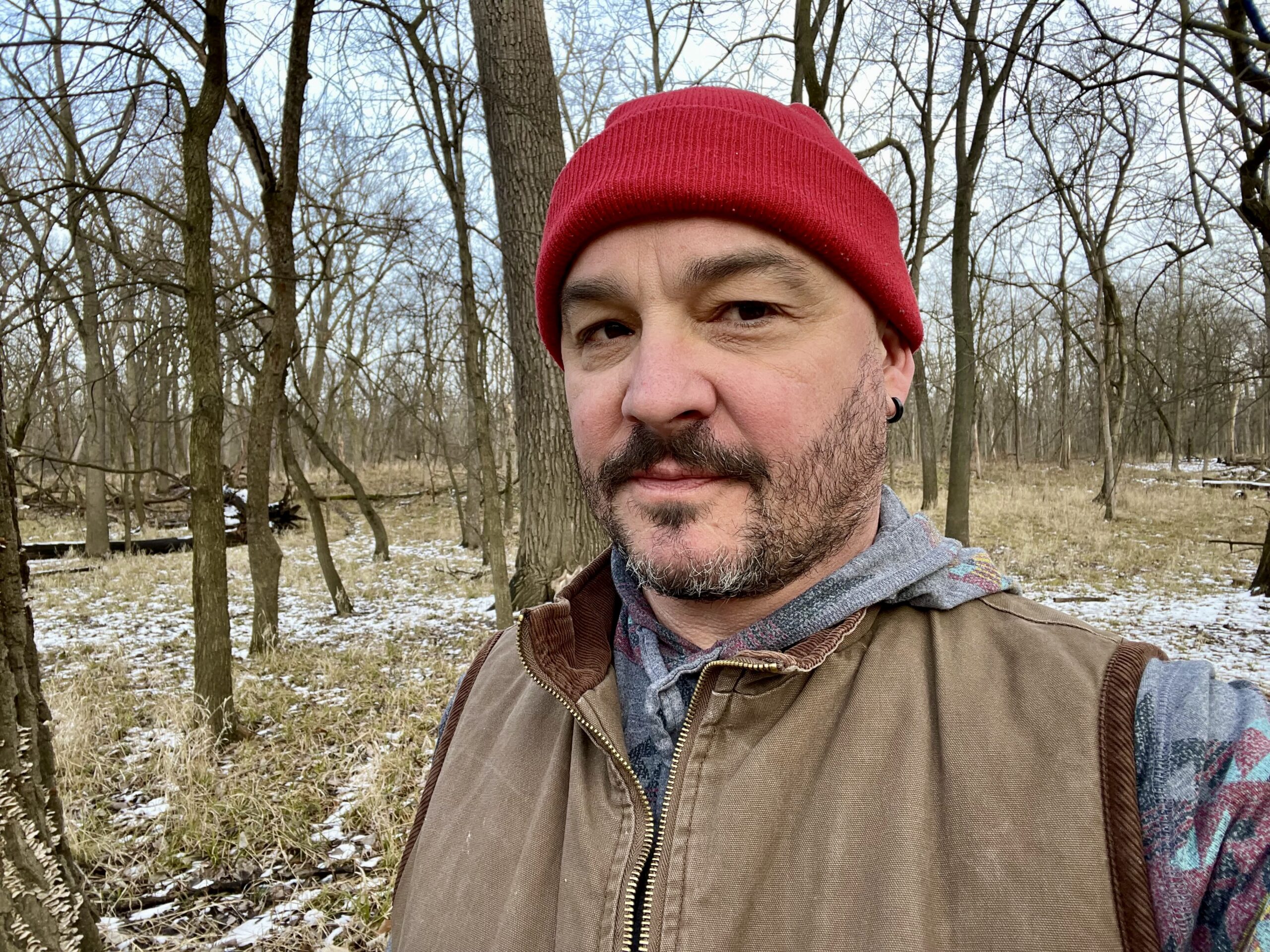 A man with beard, mustache, red hat and tan jacket looks at the camera. Bare trees of winter in the woods are behind him.