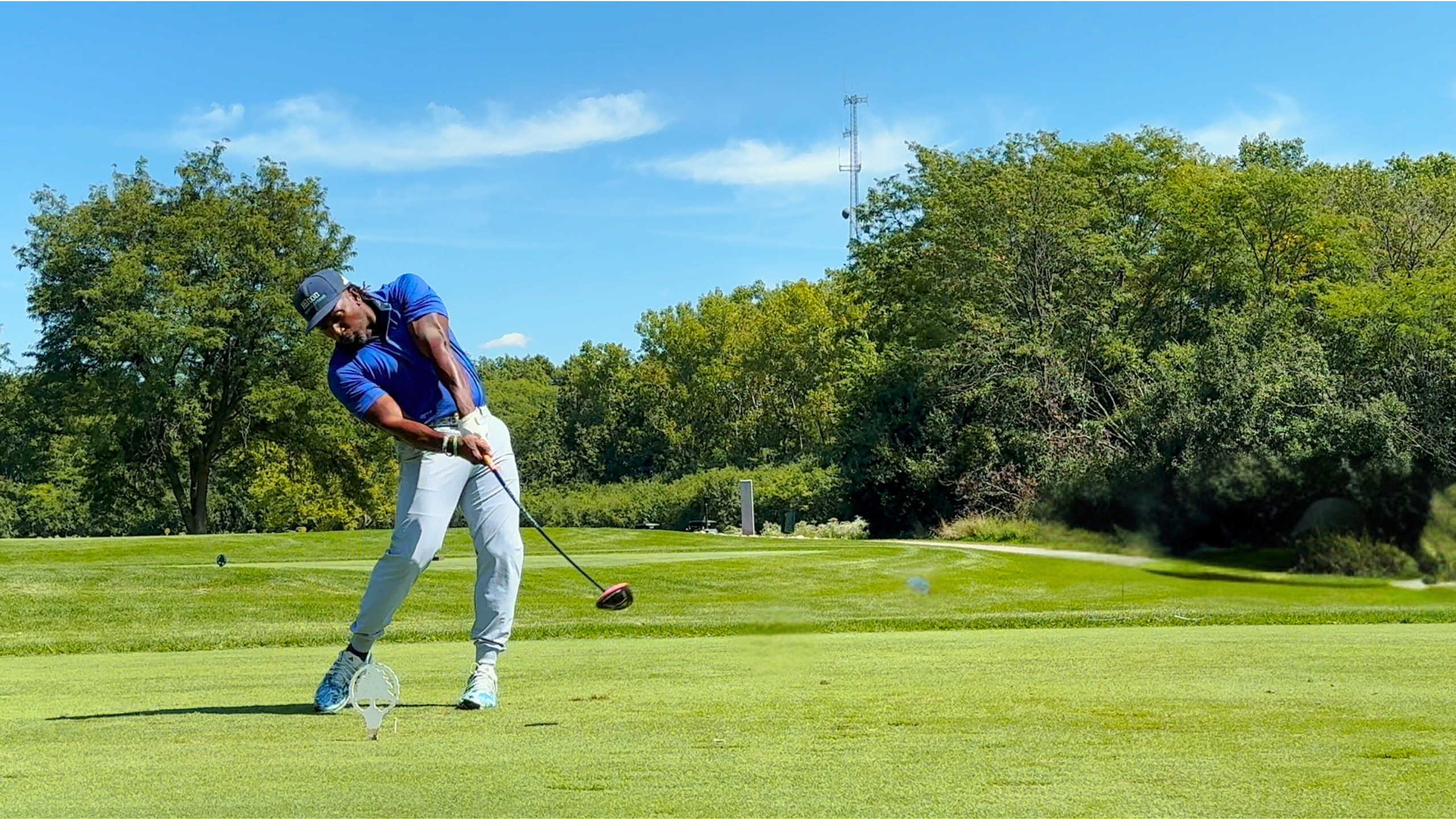 Golfer in a blue shirt and light pants on a beautiful golf course with blue sky and green trees, powerfully swinging at agolf ball