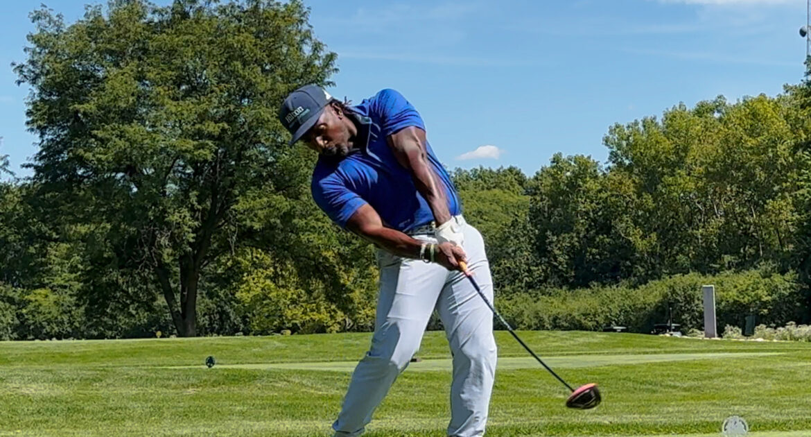 A golfer in a blue shirt and light gray pants swings at ball in a forest preserves golf course with green trees and blue sky