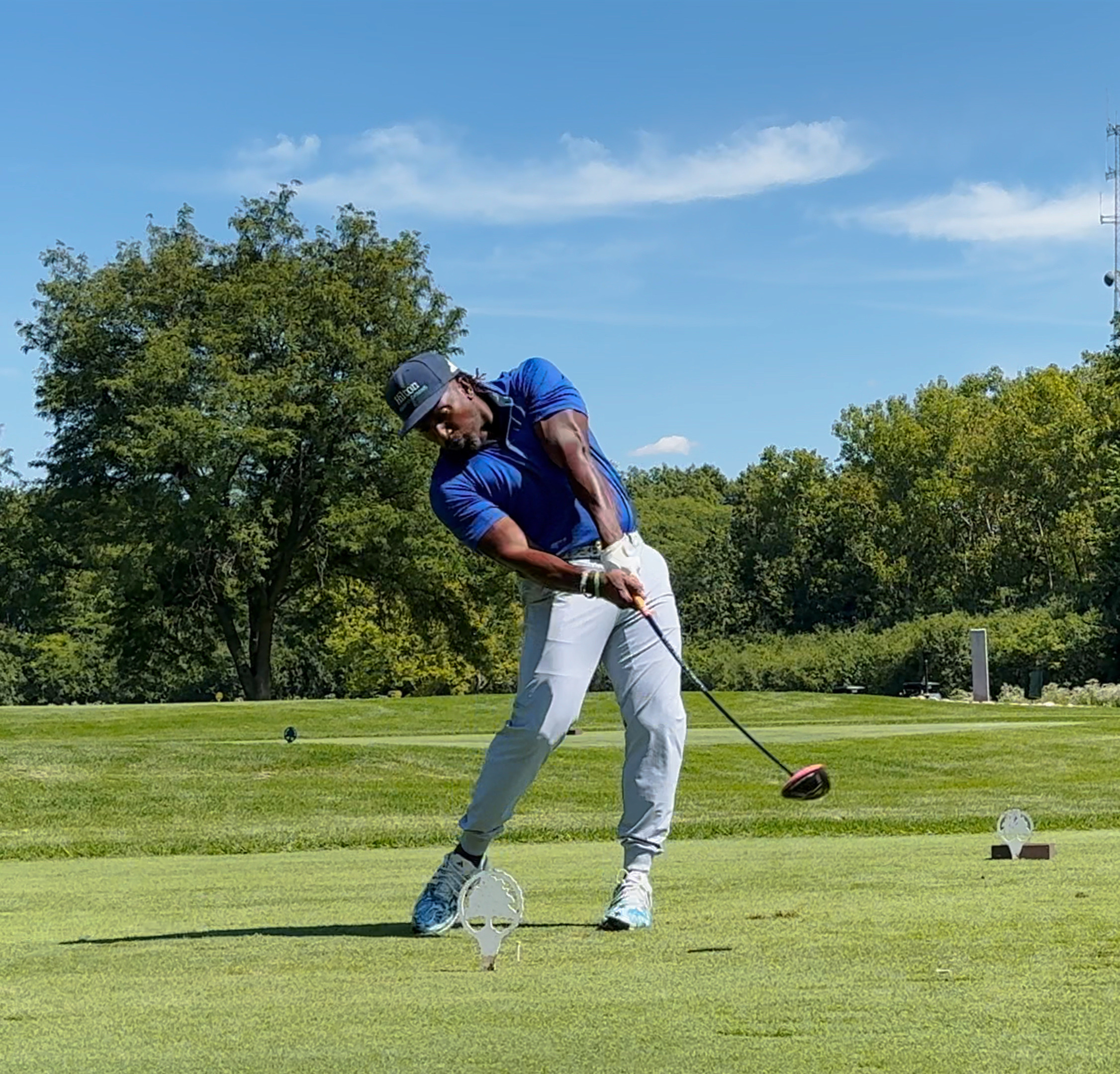 A golfer in a blue shirt and light gray pants swings at ball in a forest preserves golf course with green trees and blue sky