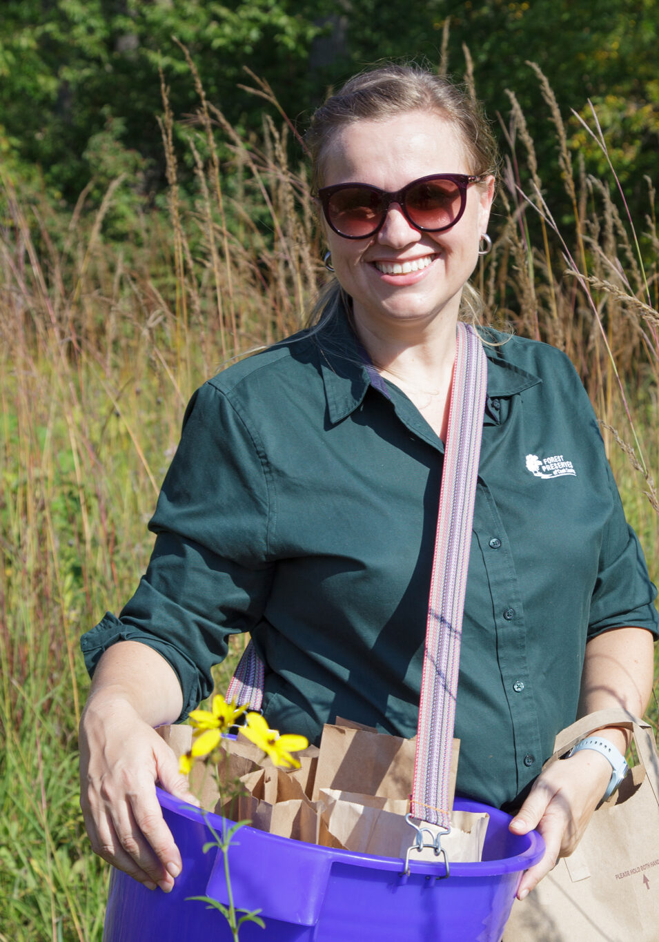 Forest Preserve employee standing in prairie in dargk green shirt and holding a purple seed harvest bucket.